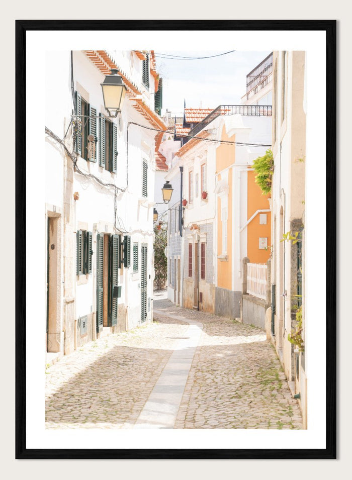 Cobbled street in a quaint European town with white buildings and a clear sky. Charming European Cobblestone Street by Henrike Schenk is a travel photography print featuring cobblestone streets, historic architecture, and European wall art.