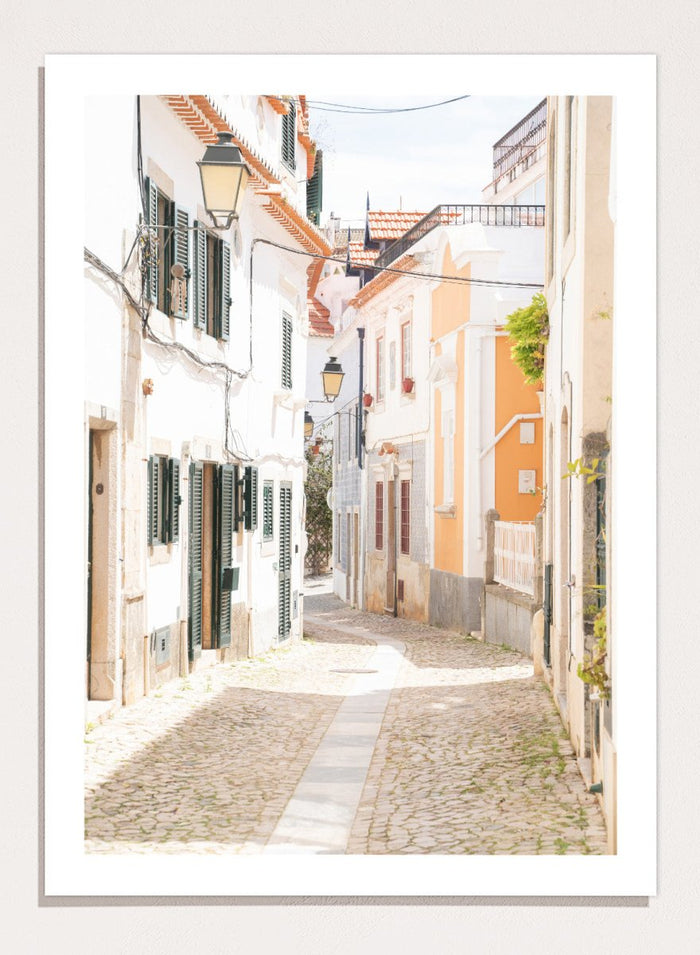 Cobbled street in a quaint European town with white buildings and a clear sky. Charming European Cobblestone Street by Henrike Schenk is a travel photography print featuring cobblestone streets, historic architecture, and European wall art.