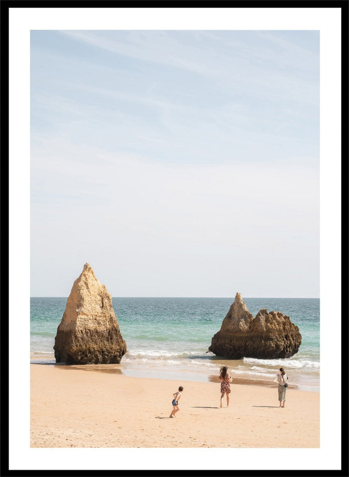 Three people on a beach with large rock formations and clear blue sky. Family Beach Day by Henrike Schenk is a coastal photography print showing a family on a sandy beach with ocean waves and dramatic sea cliffs.
