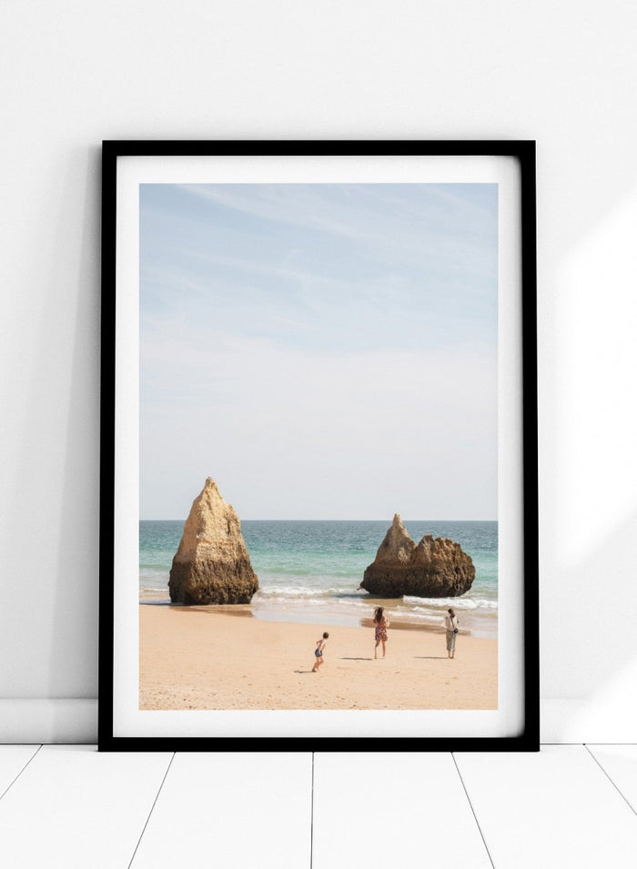 Three people on a beach with large rock formations and clear blue sky. Family Beach Day by Henrike Schenk is a coastal photography print showing a family on a sandy beach with ocean waves and dramatic sea cliffs.