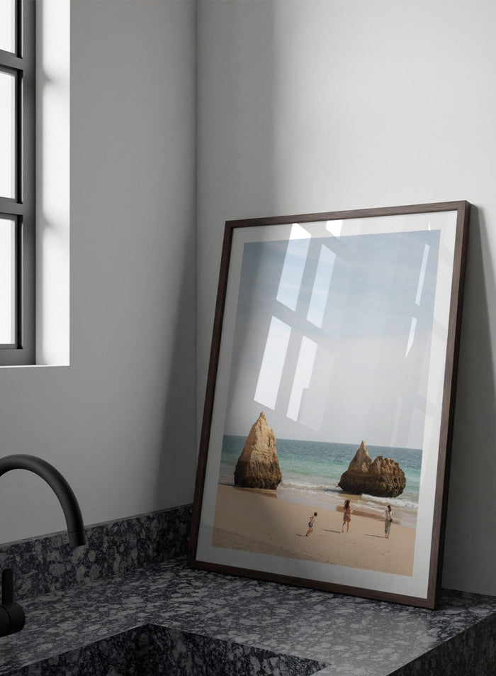 Three people on a beach with large rock formations and clear blue sky. Family Beach Day by Henrike Schenk is a coastal photography print showing a family on a sandy beach with ocean waves and dramatic sea cliffs.
