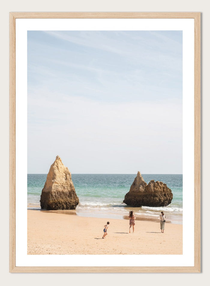 Three people on a beach with large rock formations and clear blue sky. Family Beach Day by Henrike Schenk is a coastal photography print showing a family on a sandy beach with ocean waves and dramatic sea cliffs.