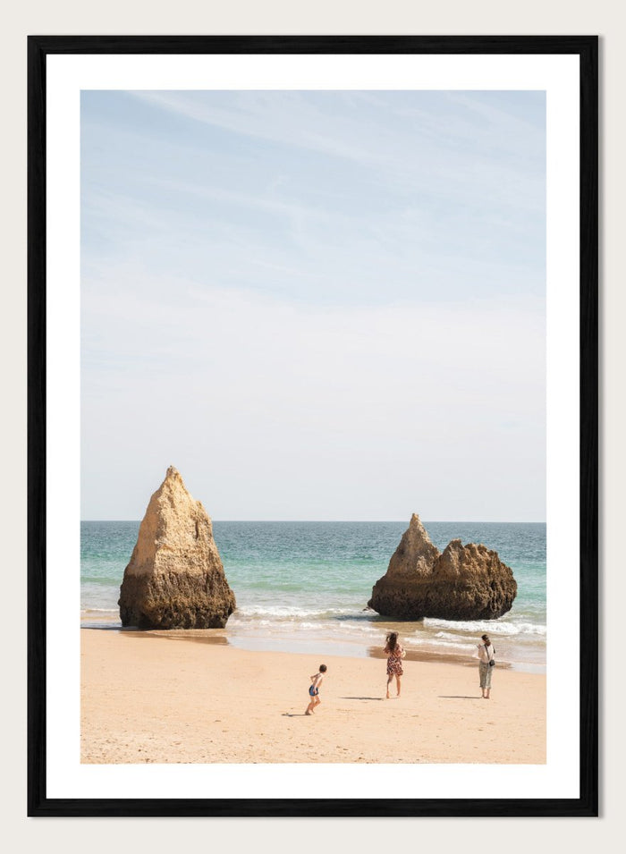 Three people on a beach with large rock formations and clear blue sky. Family Beach Day by Henrike Schenk is a coastal photography print showing a family on a sandy beach with ocean waves and dramatic sea cliffs.