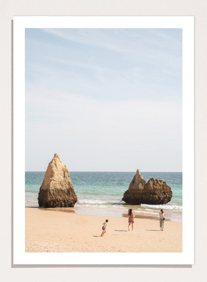Three people on a beach with large rock formations and clear blue sky. Family Beach Day by Henrike Schenk is a coastal photography print showing a family on a sandy beach with ocean waves and dramatic sea cliffs.