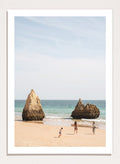 Three people on a beach with large rock formations and clear blue sky. Family Beach Day by Henrike Schenk is a coastal photography print showing a family on a sandy beach with ocean waves and dramatic sea cliffs.