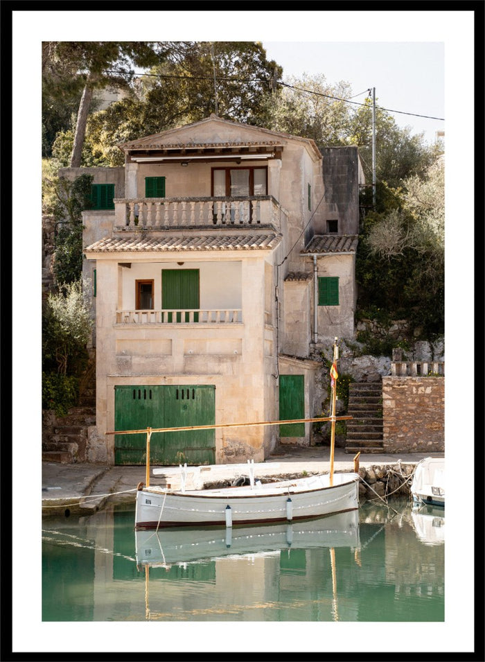 Two-story stone building with green shutters by a body of water with boats. Mediterranean Boathouse by Henrike Schenk is a coastal photography print featuring a waterfront house, fishing boat, turquoise water, and Mediterranean charm.
