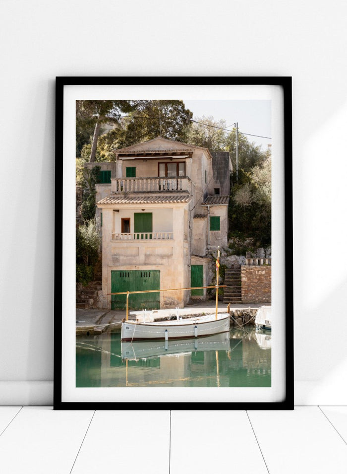 Two-story stone building with green shutters by a body of water with boats. Mediterranean Boathouse by Henrike Schenk is a coastal photography print featuring a waterfront house, fishing boat, turquoise water, and Mediterranean charm.