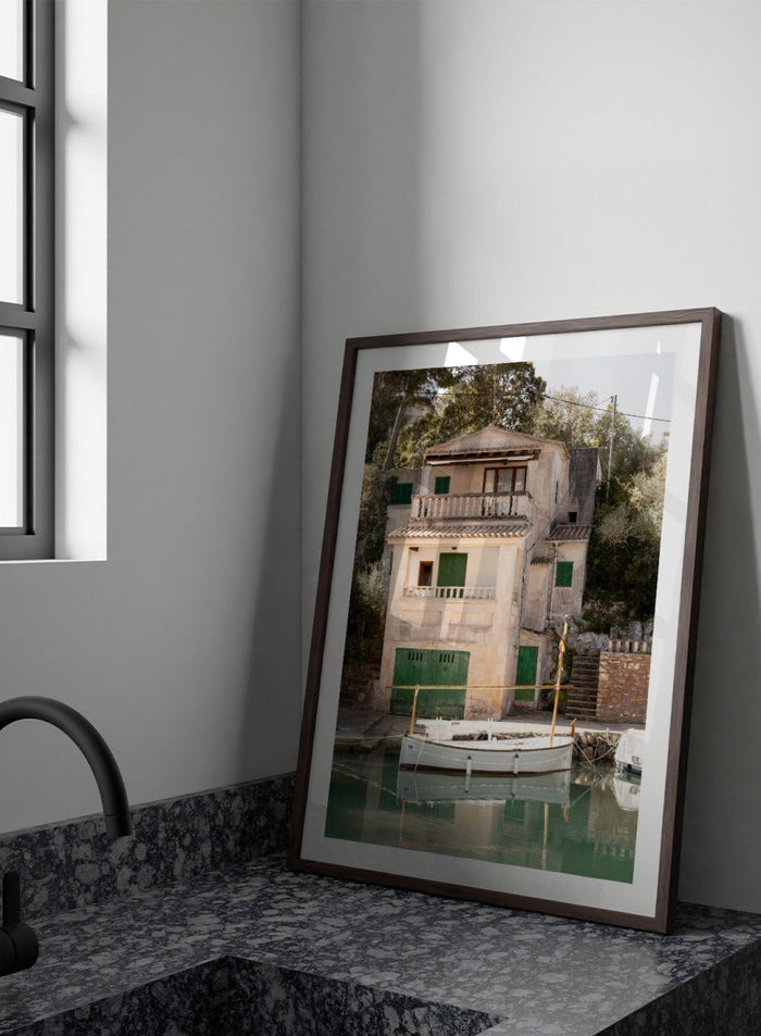 Two-story stone building with green shutters by a body of water with boats. Mediterranean Boathouse by Henrike Schenk is a coastal photography print featuring a waterfront house, fishing boat, turquoise water, and Mediterranean charm.