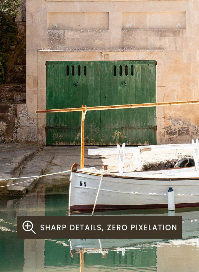 Two-story stone building with green shutters by a body of water with boats. Mediterranean Boathouse by Henrike Schenk is a coastal photography print featuring a waterfront house, fishing boat, turquoise water, and Mediterranean charm.
