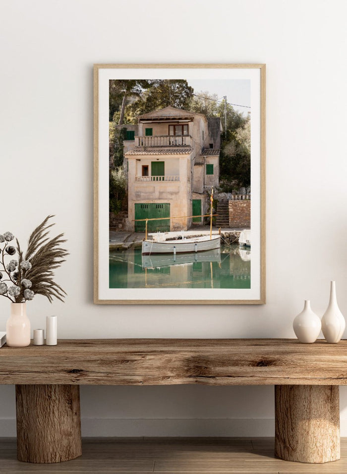 Two-story stone building with green shutters by a body of water with boats. Mediterranean Boathouse by Henrike Schenk is a coastal photography print featuring a waterfront house, fishing boat, turquoise water, and Mediterranean charm.