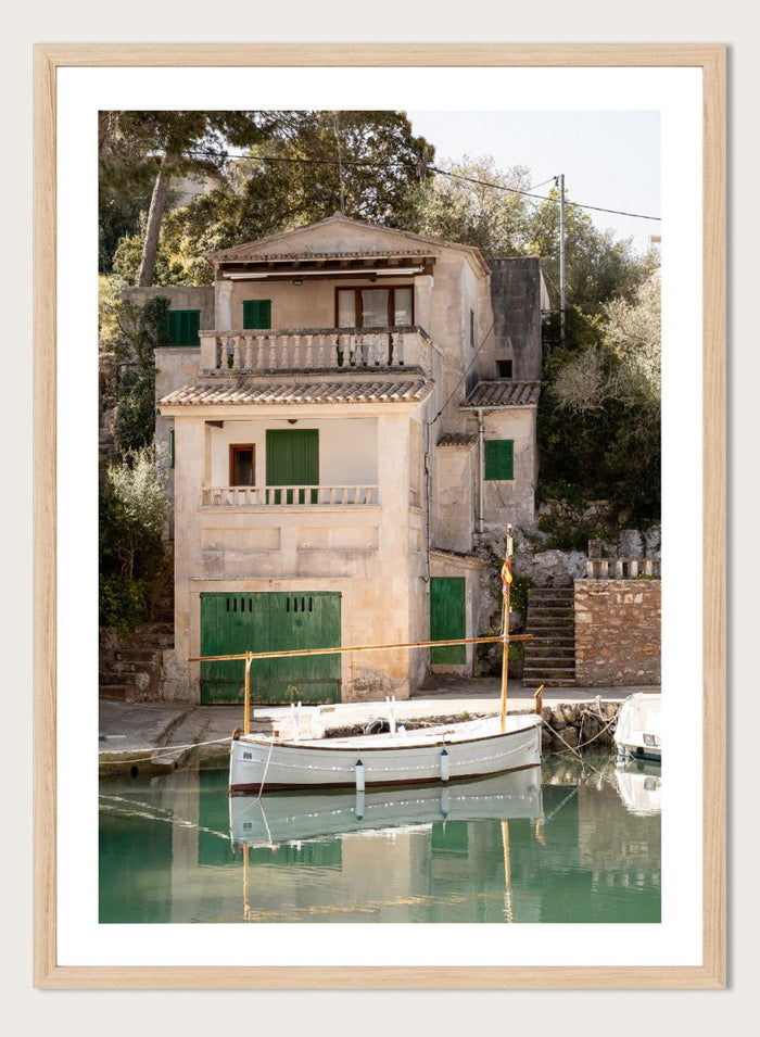 Two-story stone building with green shutters by a body of water with boats. Mediterranean Boathouse by Henrike Schenk is a coastal photography print featuring a waterfront house, fishing boat, turquoise water, and Mediterranean charm.