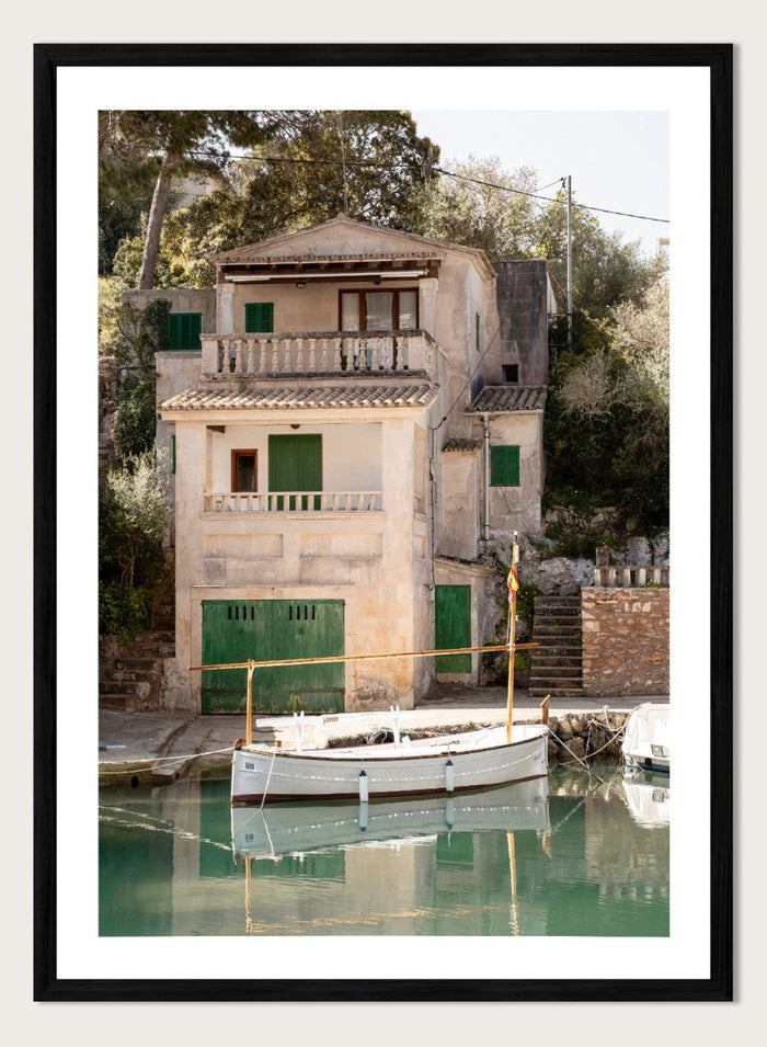 Two-story stone building with green shutters by a body of water with boats. Mediterranean Boathouse by Henrike Schenk is a coastal photography print featuring a waterfront house, fishing boat, turquoise water, and Mediterranean charm.