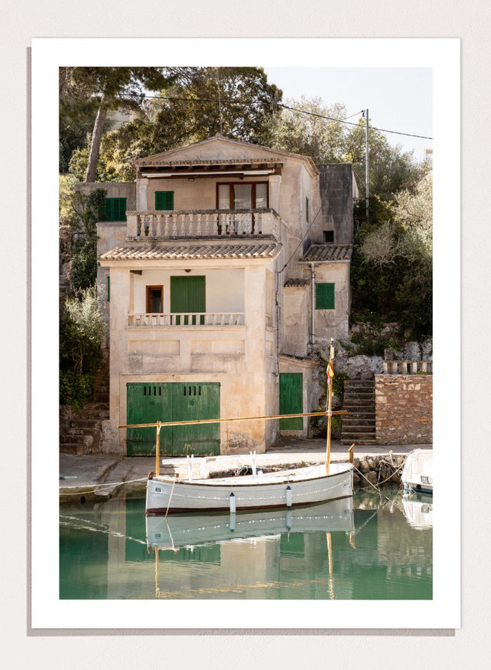 Two-story stone building with green shutters by a body of water with boats. Mediterranean Boathouse by Henrike Schenk is a coastal photography print featuring a waterfront house, fishing boat, turquoise water, and Mediterranean charm.