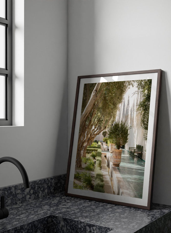 Garden pathway with trees, plants, and a building in the background. Mediterranean Garden Courtyard by Henrike Schenk is a travel photography print featuring olive trees, terracotta pots, tiled walkways, and a Moroccan garden.