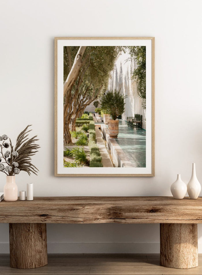 Garden pathway with trees, plants, and a building in the background. Mediterranean Garden Courtyard by Henrike Schenk is a travel photography print featuring olive trees, terracotta pots, tiled walkways, and a Moroccan garden.