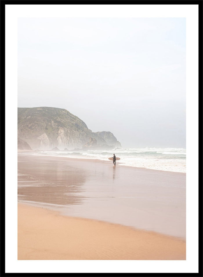 Person walking along a beach with mountains in the background. Misty Morning Surfer by Henrike Schenk captures a surfer walking along a misty beach with ocean waves, coastal scenery, and photography wall art.