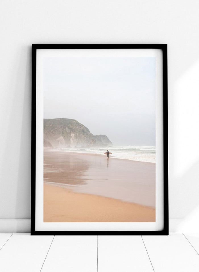 Person walking along a beach with mountains in the background. Misty Morning Surfer by Henrike Schenk captures a surfer walking along a misty beach with ocean waves, coastal scenery, and photography wall art.