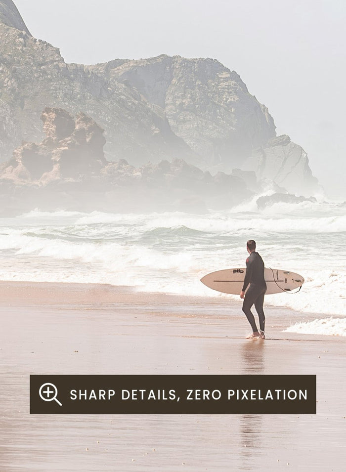 Person walking along a beach with mountains in the background. Misty Morning Surfer by Henrike Schenk captures a surfer walking along a misty beach with ocean waves, coastal scenery, and photography wall art.