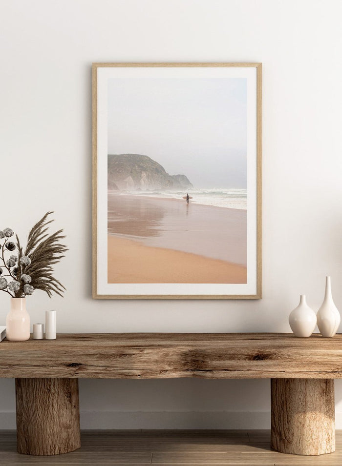 Person walking along a beach with mountains in the background. Misty Morning Surfer by Henrike Schenk captures a surfer walking along a misty beach with ocean waves, coastal scenery, and photography wall art.