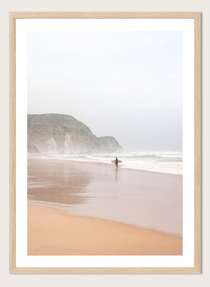 Person walking along a beach with mountains in the background. Misty Morning Surfer by Henrike Schenk captures a surfer walking along a misty beach with ocean waves, coastal scenery, and photography wall art.