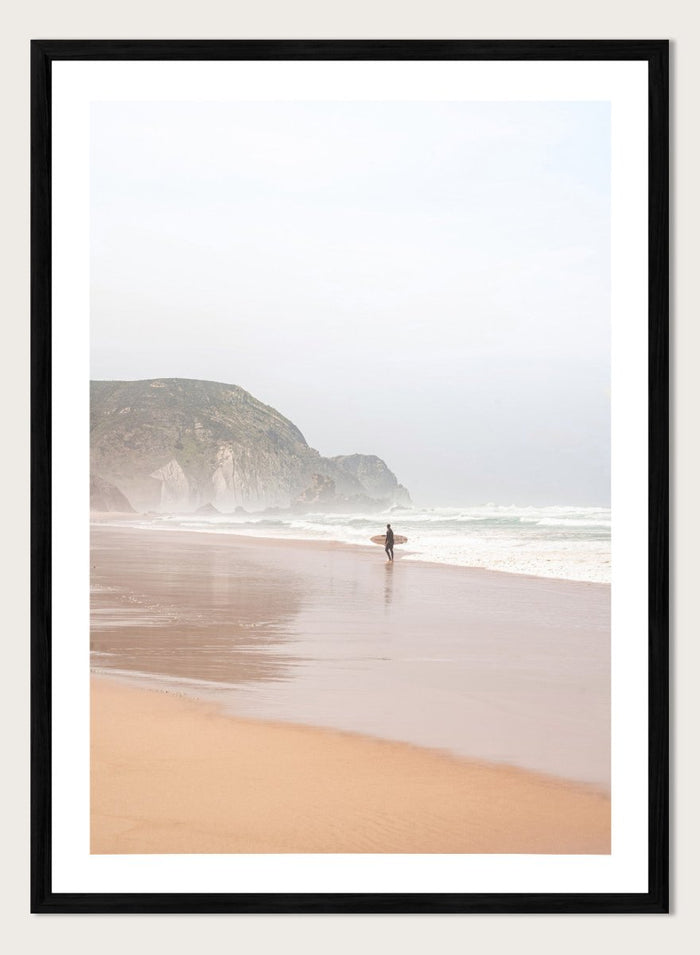 Person walking along a beach with mountains in the background. Misty Morning Surfer by Henrike Schenk captures a surfer walking along a misty beach with ocean waves, coastal scenery, and photography wall art.