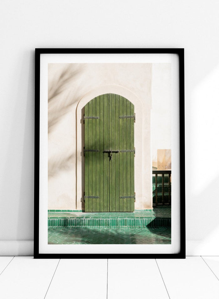 Green wooden door with metal latch on a white wall next to a pool. Moroccan Green Door by Henrike Schenk is an architectural photography print featuring green tilework, a traditional doorway, and Moroccan design details.