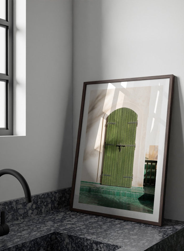 Green wooden door with metal latch on a white wall next to a pool. Moroccan Green Door by Henrike Schenk is an architectural photography print featuring green tilework, a traditional doorway, and Moroccan design details.