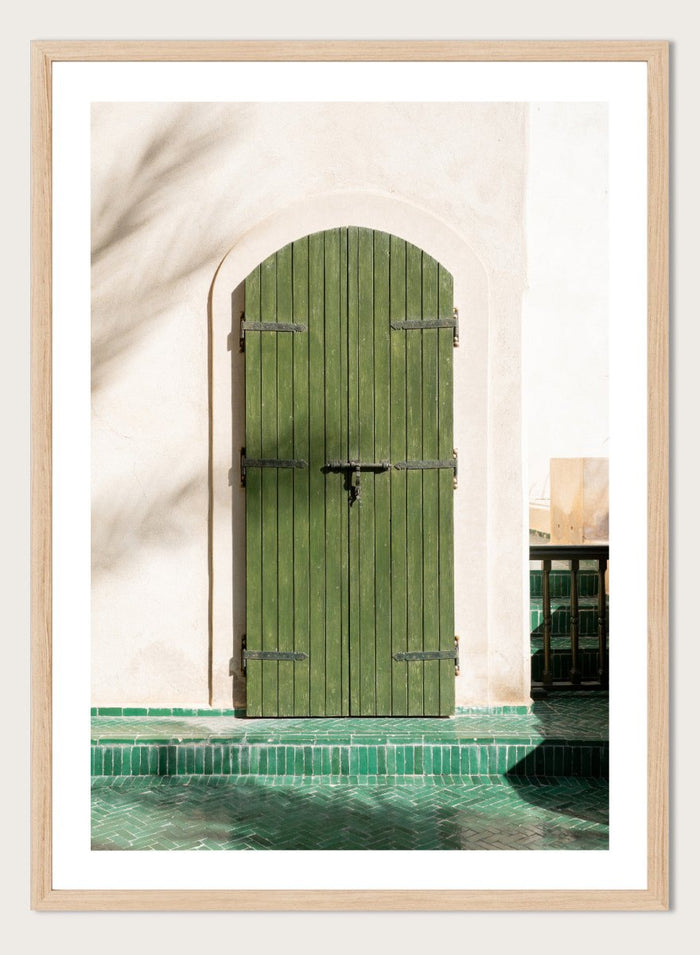Green wooden door with metal latch on a white wall next to a pool. Moroccan Green Door by Henrike Schenk is an architectural photography print featuring green tilework, a traditional doorway, and Moroccan design details.