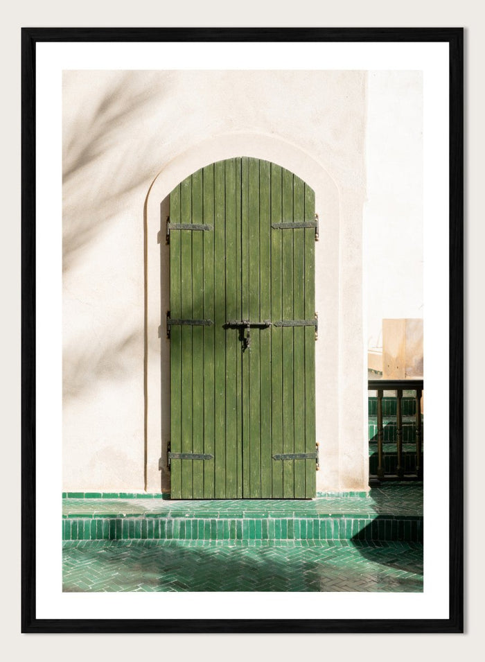 Green wooden door with metal latch on a white wall next to a pool. Moroccan Green Door by Henrike Schenk is an architectural photography print featuring green tilework, a traditional doorway, and Moroccan design details.
