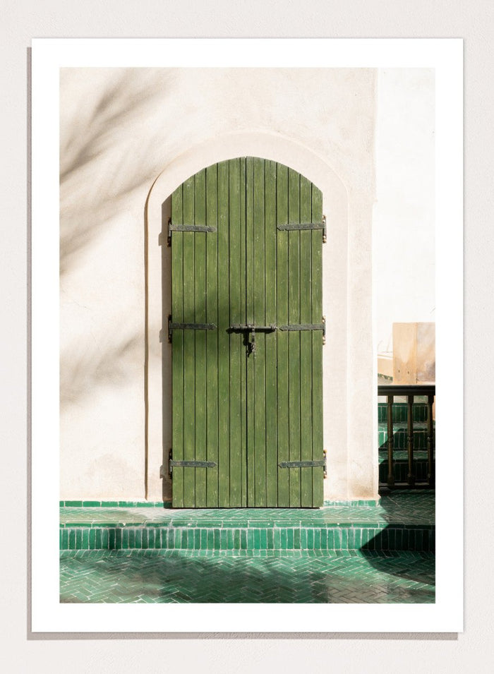 Green wooden door with metal latch on a white wall next to a pool. Moroccan Green Door by Henrike Schenk is an architectural photography print featuring green tilework, a traditional doorway, and Moroccan design details.