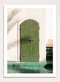 Green wooden door with metal latch on a white wall next to a pool. Moroccan Green Door by Henrike Schenk is an architectural photography print featuring green tilework, a traditional doorway, and Moroccan design details.
