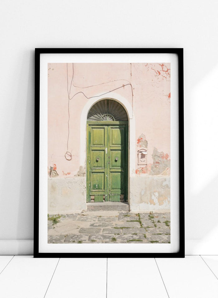 Green door on a pink wall with peeling paint. Old World Green Door by Henrike Schenk features a vintage European doorway, weathered architecture, pastel walls, travel photography, and wall art.