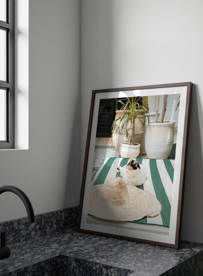 Cat lying on a green and white striped towel with a straw hat nearby, potted plants in the background. Patio Cat and Straw Hat by Henrike Schenk is a Mediterranean photography print featuring a cat, striped lounge chair, courtyard decor, and summer travel style.