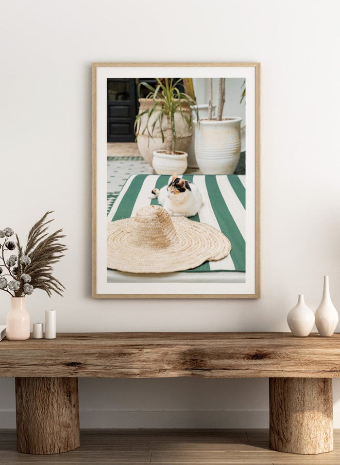 Cat lying on a green and white striped towel with a straw hat nearby, potted plants in the background. Patio Cat and Straw Hat by Henrike Schenk is a Mediterranean photography print featuring a cat, striped lounge chair, courtyard decor, and summer travel style.