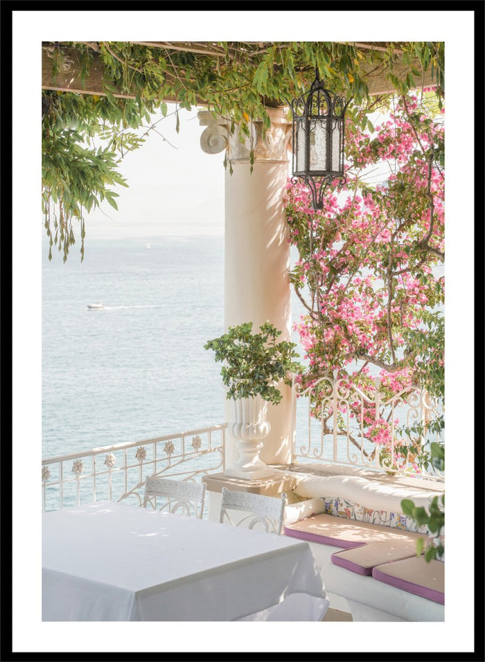Outdoor seating area with flowers and ocean view. Pink Flowers Ocean View by Henrike Schenk is a coastal photography print featuring blooming flowers, Mediterranean architecture, ocean views, and soft natural light.