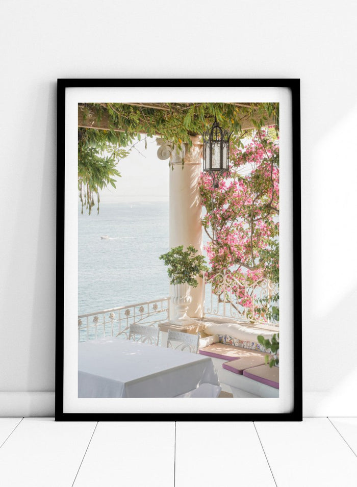 Outdoor seating area with flowers and ocean view. Pink Flowers Ocean View by Henrike Schenk is a coastal photography print featuring blooming flowers, Mediterranean architecture, ocean views, and soft natural light.