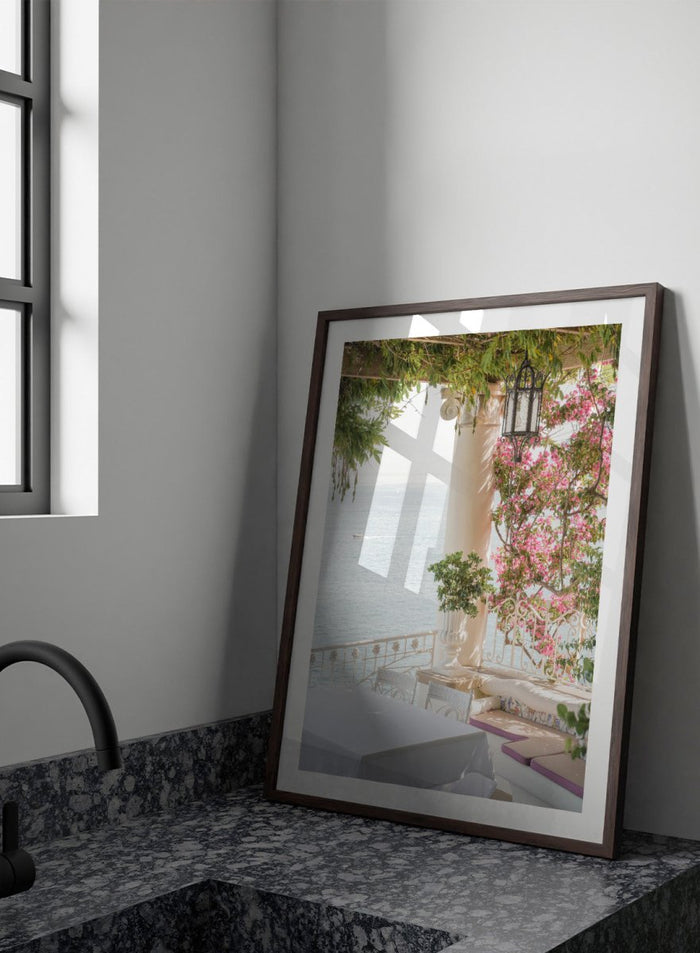 Outdoor seating area with flowers and ocean view. Pink Flowers Ocean View by Henrike Schenk is a coastal photography print featuring blooming flowers, Mediterranean architecture, ocean views, and soft natural light.