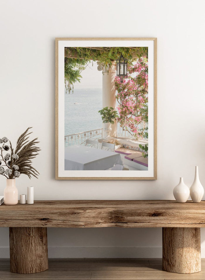 Outdoor seating area with flowers and ocean view. Pink Flowers Ocean View by Henrike Schenk is a coastal photography print featuring blooming flowers, Mediterranean architecture, ocean views, and soft natural light.