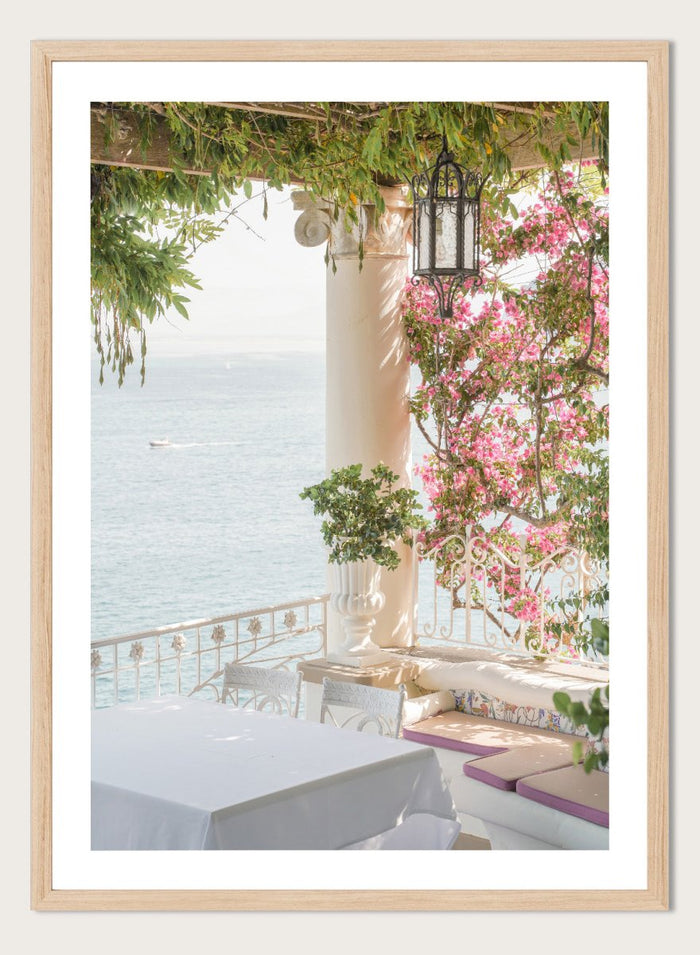 Outdoor seating area with flowers and ocean view. Pink Flowers Ocean View by Henrike Schenk is a coastal photography print featuring blooming flowers, Mediterranean architecture, ocean views, and soft natural light.