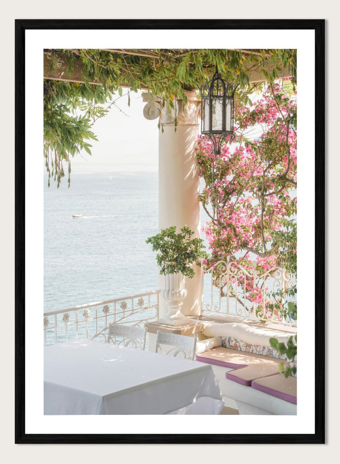 Outdoor seating area with flowers and ocean view. Pink Flowers Ocean View by Henrike Schenk is a coastal photography print featuring blooming flowers, Mediterranean architecture, ocean views, and soft natural light.