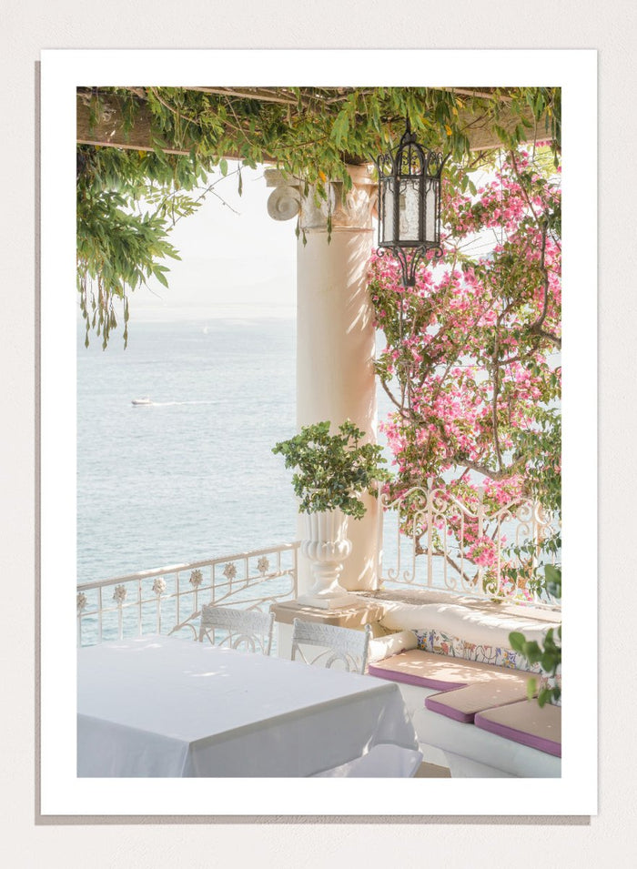 Outdoor seating area with flowers and ocean view. Pink Flowers Ocean View by Henrike Schenk is a coastal photography print featuring blooming flowers, Mediterranean architecture, ocean views, and soft natural light.