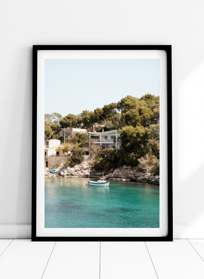Cottage by a lake with trees and boats in the water. Serene Mediterranean Cove by Henrike Schenk is a coastal photography print featuring turquoise water, a fishing boat, seaside homes, and Mediterranean scenery.