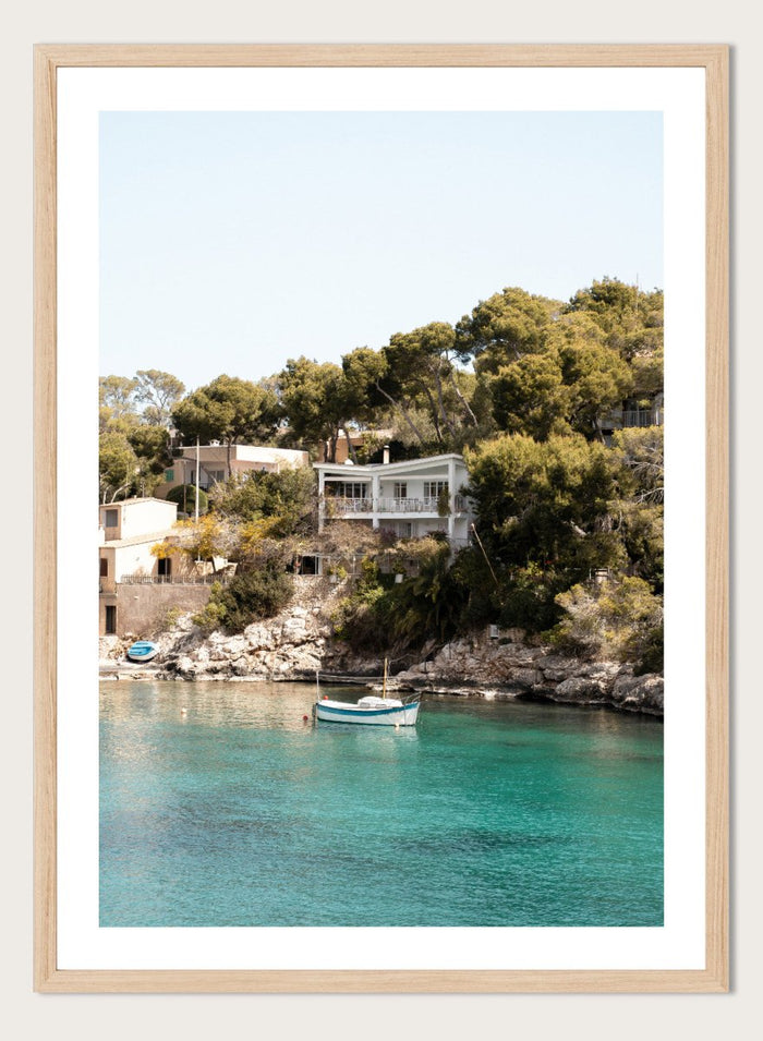 Cottage by a lake with trees and boats in the water. Serene Mediterranean Cove by Henrike Schenk is a coastal photography print featuring turquoise water, a fishing boat, seaside homes, and Mediterranean scenery.