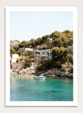 Cottage by a lake with trees and boats in the water. Serene Mediterranean Cove by Henrike Schenk is a coastal photography print featuring turquoise water, a fishing boat, seaside homes, and Mediterranean scenery.