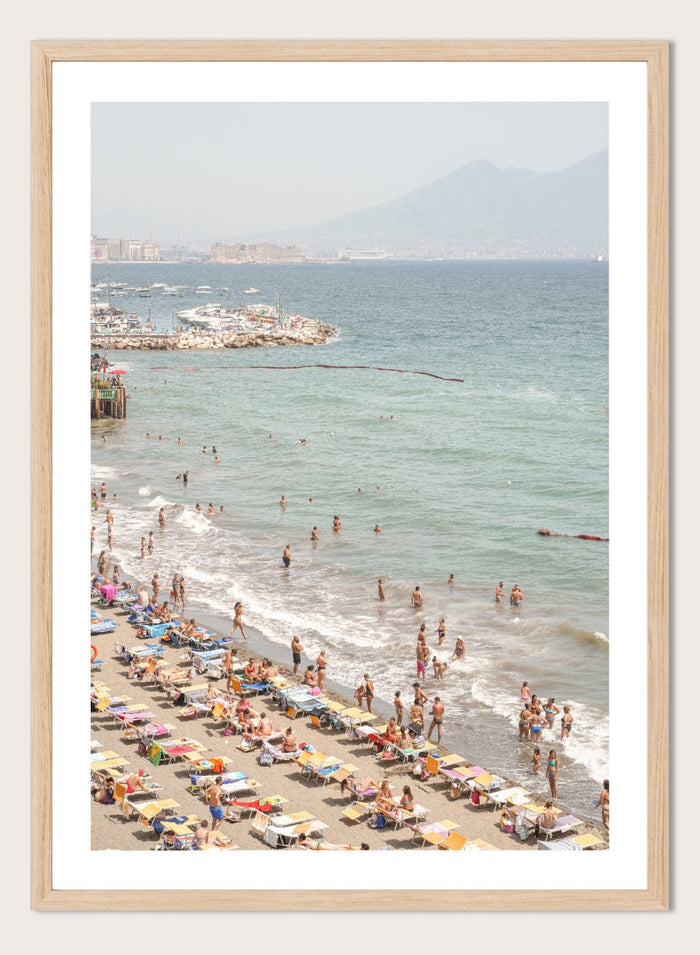 Beach scene with people and umbrellas near a body of water with mountains in the background. Summer Beach Mountain View by Henrike Schenk is a Mediterranean beach photography print featuring coastal views, mountains, ocean scenery, and summer travel.