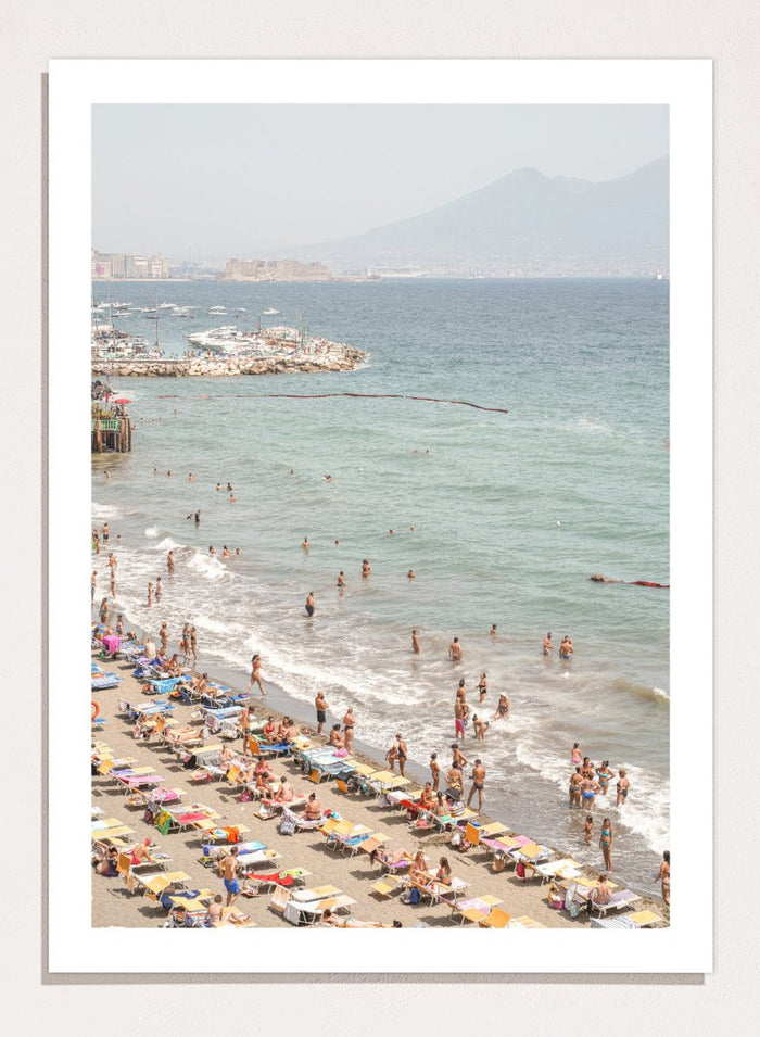 Beach scene with people and umbrellas near a body of water with mountains in the background. Summer Beach Mountain View by Henrike Schenk is a Mediterranean beach photography print featuring coastal views, mountains, ocean scenery, and summer travel.