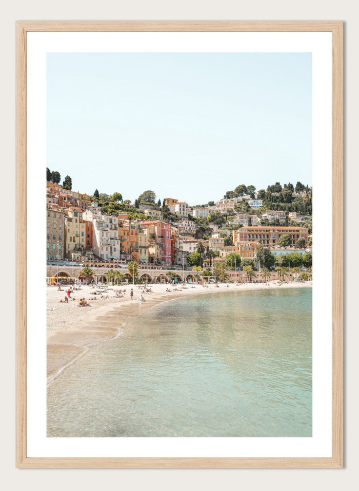 Coastal town with colorful buildings and a beach. Sunny Mediterranean Coast by Henrike Schenk is a coastal photography print featuring turquoise water, pastel buildings, Mediterranean beach scenery, and travel wall art.