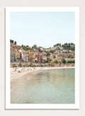 Coastal town with colorful buildings and a beach. Sunny Mediterranean Coast by Henrike Schenk is a coastal photography print featuring turquoise water, pastel buildings, Mediterranean beach scenery, and travel wall art.