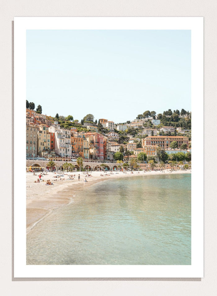 Coastal town with colorful buildings and a beach. Sunny Mediterranean Coast by Henrike Schenk is a coastal photography print featuring turquoise water, pastel buildings, Mediterranean beach scenery, and travel wall art.