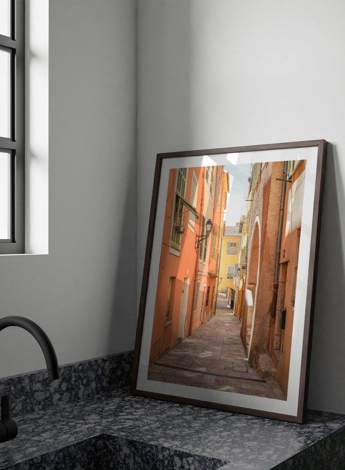 Framed photograph of a narrow street on a wall next to a window.  narrow street with colorful buildings in a European city. Vibrant European Alley by Henrike Schenk is a travel photography print featuring a Mediterranean street, cobblestone alley, and pastel European architecture.